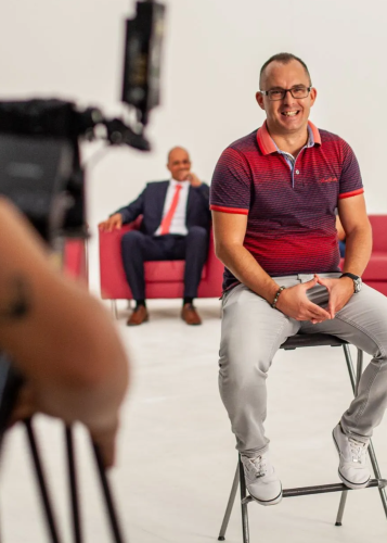 Két férfi egy forgatáson vagy interjúhelyzetben, fehér háttérrel és rózsaszín kanapéval. Az egyik férfi előtérben mosolyogva ül egy széken, a másik háttérben elegáns öltönyben, lazán ül a kanapén. Kamera, interjú, forgatás, üzlet, kommunikáció, média, stú