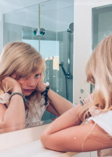 Egy fiatal szőke lány tükörképét mutatja a fürdőszobában, aki láthatóan szomorú vagy gondterhelt. A lány alkarjára támasztja az állát, szemei lefelé néznek, arca kifejezéstelen. Hosszú, szőke haja az arcába lóg.