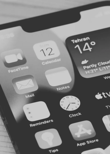 Fekete-fehér kép egy okostelefon képernyőjéről, melyen különböző alkalmazásikonok láthatók, mint például FaceTime, Calendar, Mail, Notes, Clock, Reminders, App Store és egy időjárás widget. Okostelefon, mobilalkalmazások, digitális eszköz, felhasználói fe