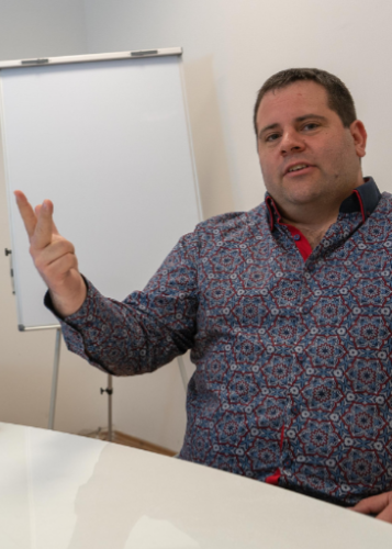 Egy férfi ül egy asztalnál, elegáns, mintás ingben, kezével magyarázó mozdulatot tesz, háttérben egy üres flipchart tábla. Fókuszált tekintettel beszél. Prezentáció, megbeszélés, üzleti találkozó, magyarázat, szakértő, oktatás, kommunikáció, irodai környe