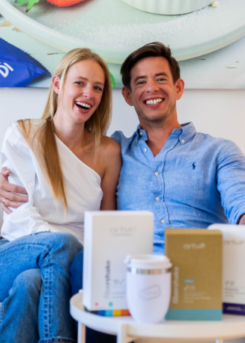 Egy boldog, mosolygós pár ül egy kanapén, és a kamerába néz, miközben az asztalon fehér és arany színű dobozok (amelyek a "natrue" márkajelzést viselik, az egyik "shakes", a másik "electrolytes" felirattal), valamint egy fehér termoszkupa látható.