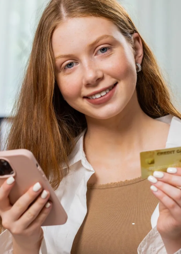 Közelkép egy mosolygós, fiatal nőről, aki telefonon keresztül online vásárol vagy banki ügyeket intéz. A vörös hajú, világos bőrű nő a kamerába néz és mosolyog. Jobb kezében egy rózsaszín mobiltelefont tart, bal kezében pedig egy arany színű bankkártyát.