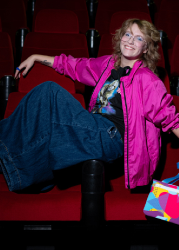 Mosolygó fiatal nő szemüvegben, neonrózsaszín dzsekiben és bő farmernadrágban pózol egy üres, vörös székkel teli moziteremben. Mozi, szórakozás, divat, stílus, fiatalos és bohókás portré, szabadidő, kulturális program.